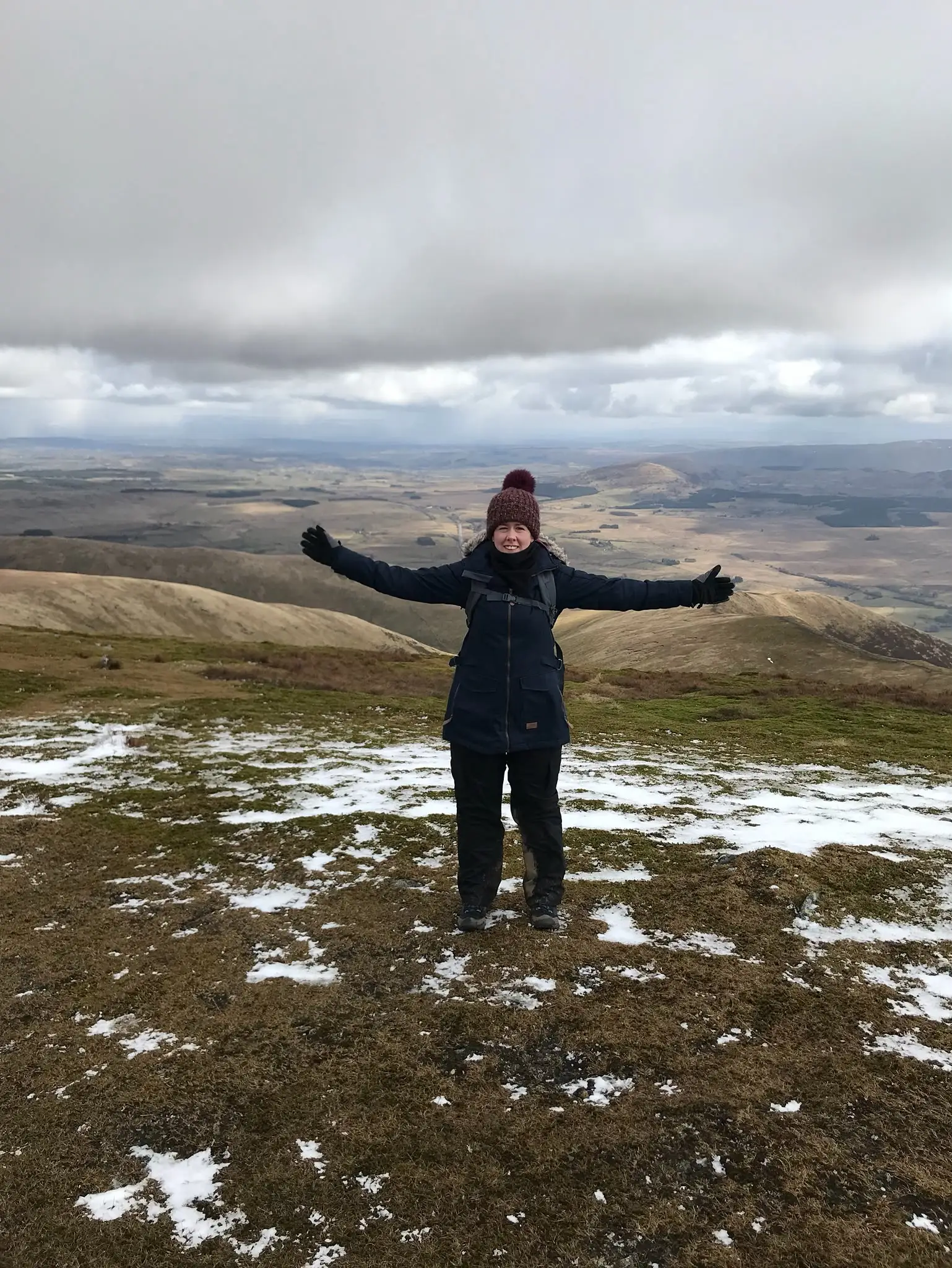 Laura Heslop standing on a hilltop with arms spread wide, smiling, snow patches on the ground and rolling countryside behind her.