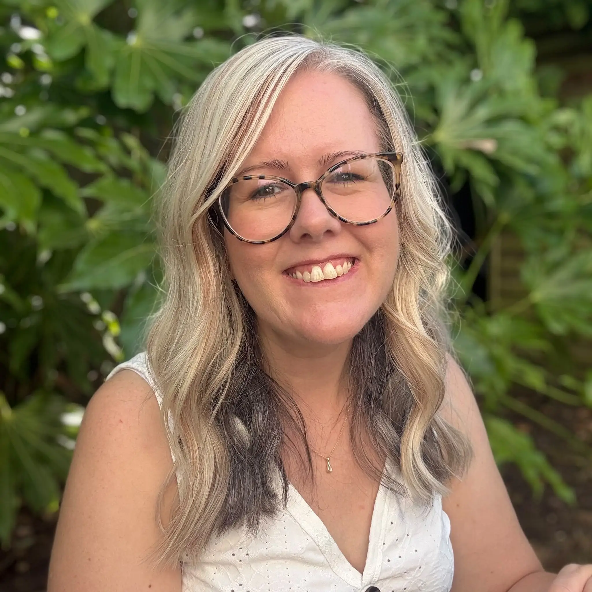 Laura Heslop, therapist, smiling to camera in a white top with long blonde hair and glasses, framed by green foliage.
