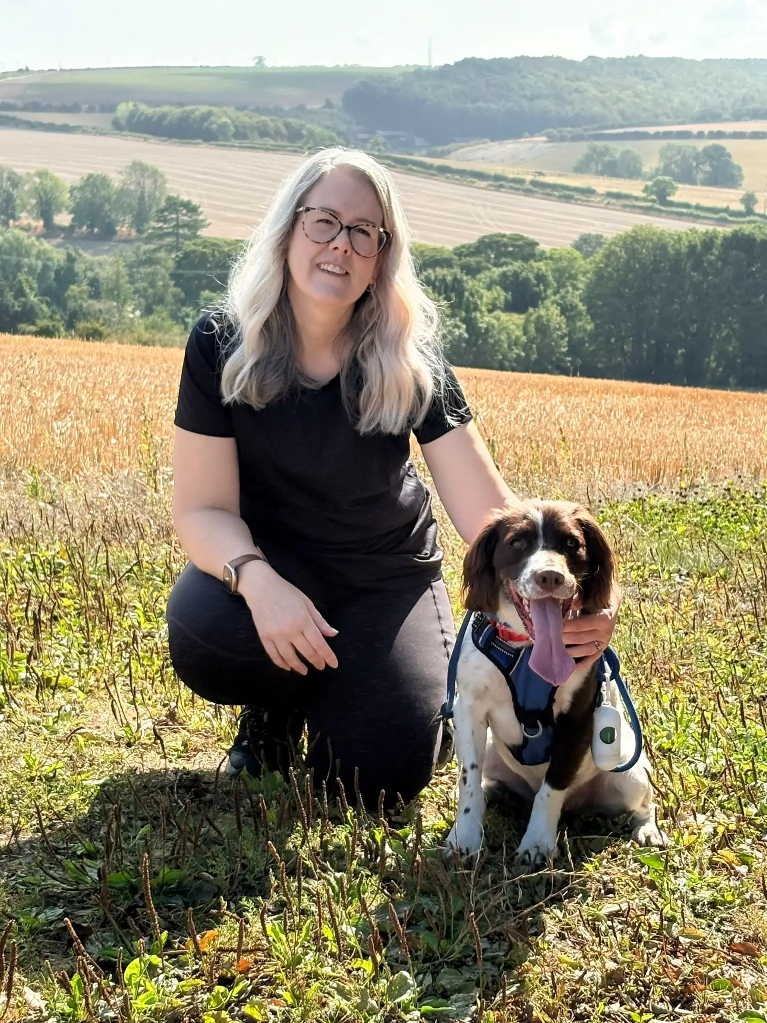 Laura Heslop kneeling in a sunlit field beside a springer spaniel with its tongue out, countryside behind them.